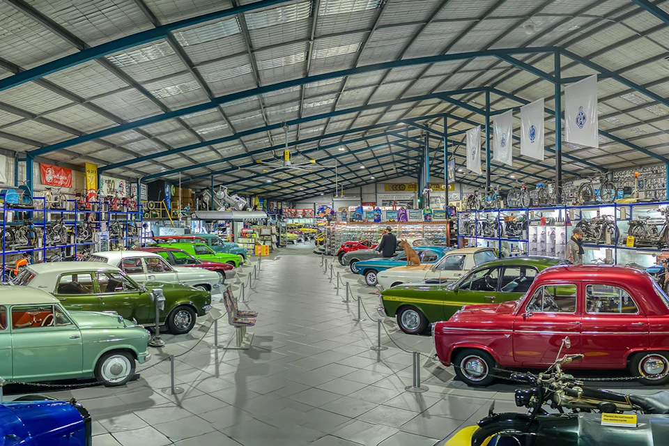 A spacious indoor vintage car museum displays rows of colorful classic cars under a large, high-ceilinged metal roof.