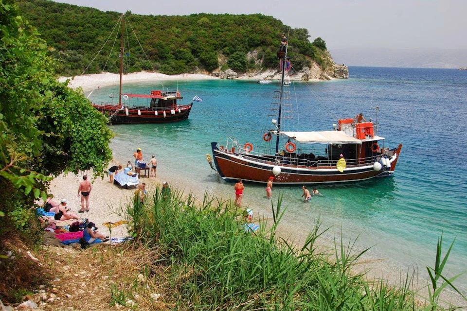 People enjoying a beach with two docked wooden boats in clear blue water, surrounded by vegetation and a forested background.