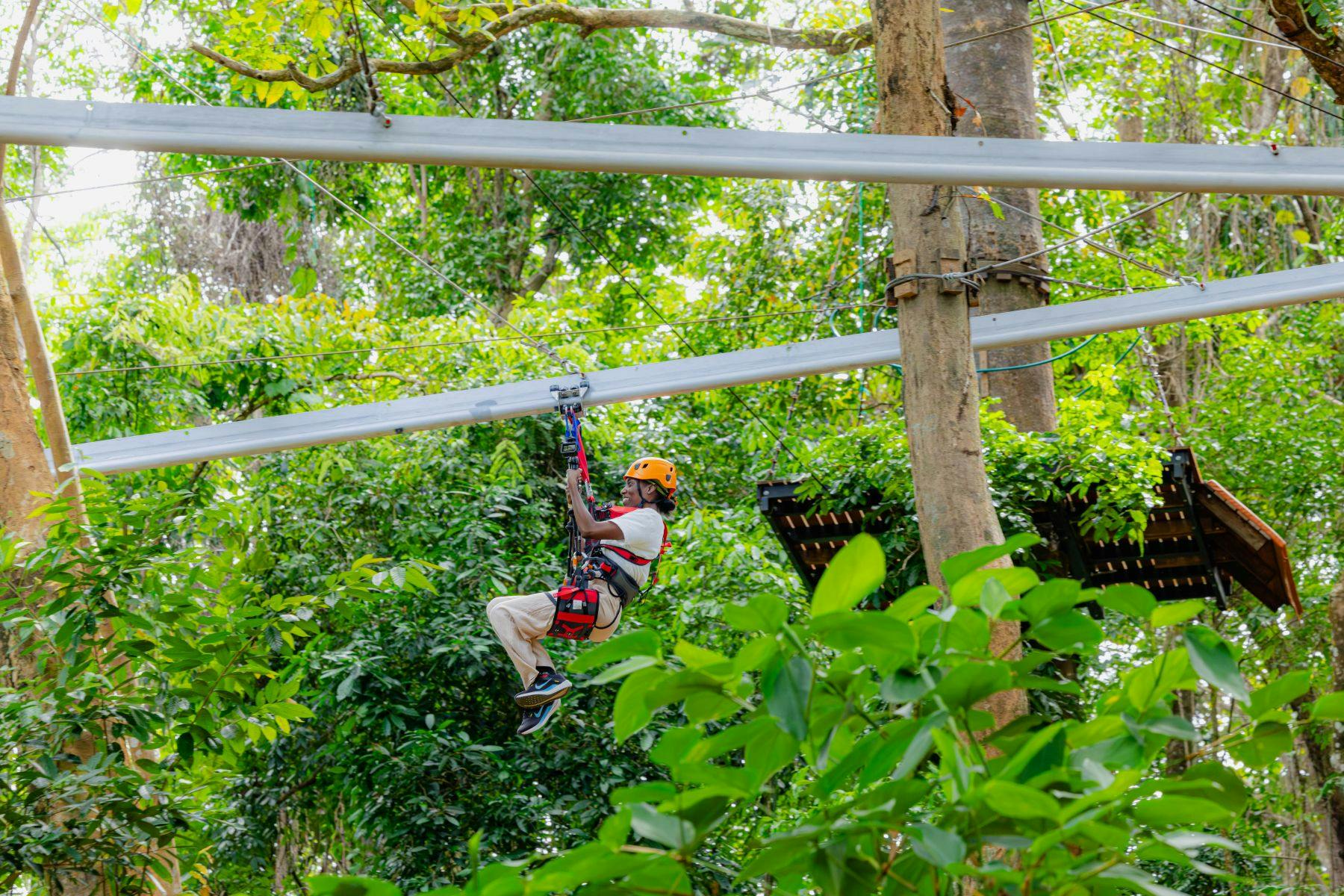 A person wearing a harness and helmet ziplining through a lush green forest on a cable system.