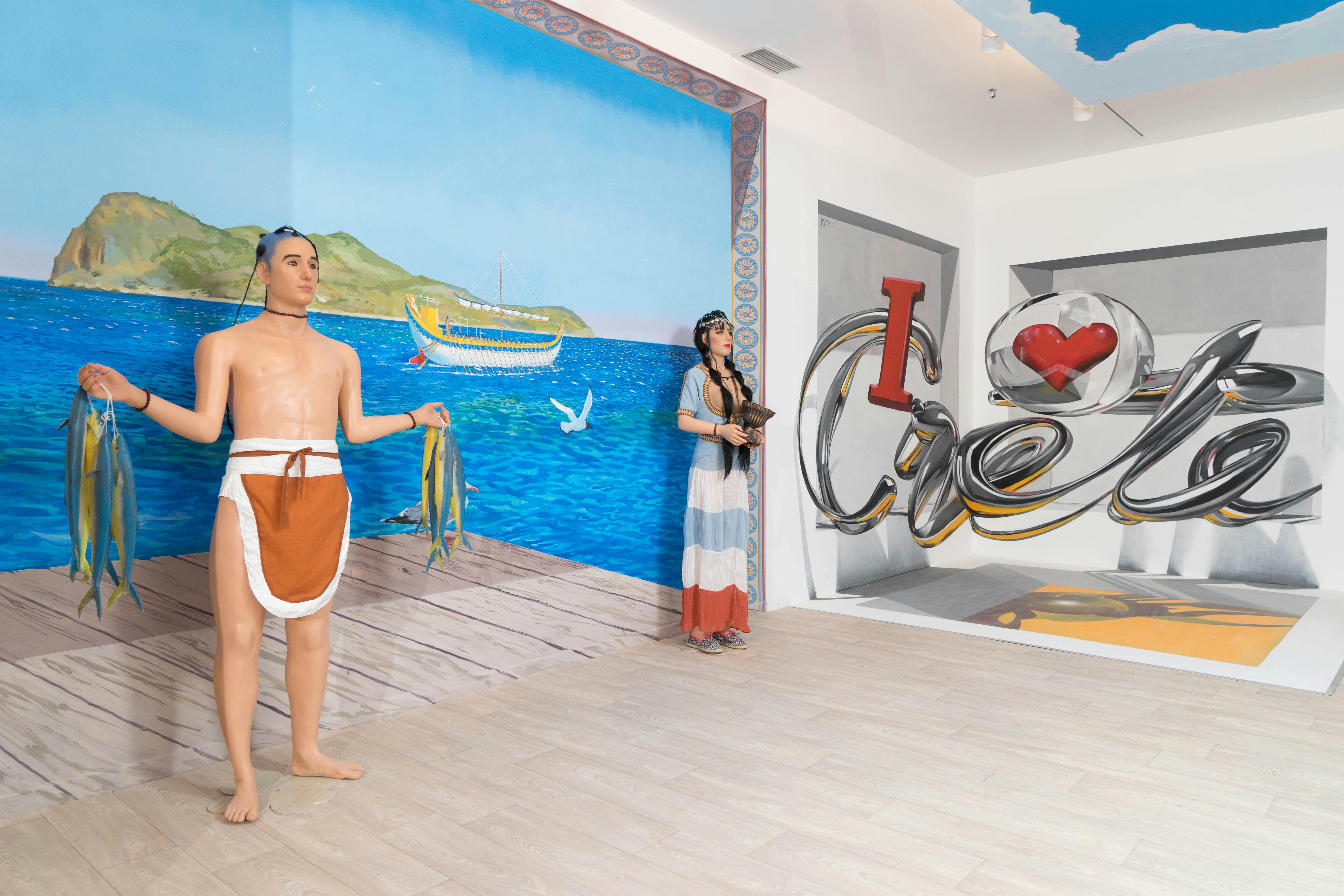 Dvě figuríny ve starobylém oděvu stojí na doku s nástěnnou malbou pobřežní scény a lodí v pozadí.