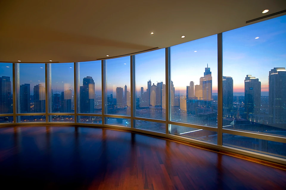 Spacious room with floor-to-ceiling windows offering a panoramic view of a city skyline at sunset.