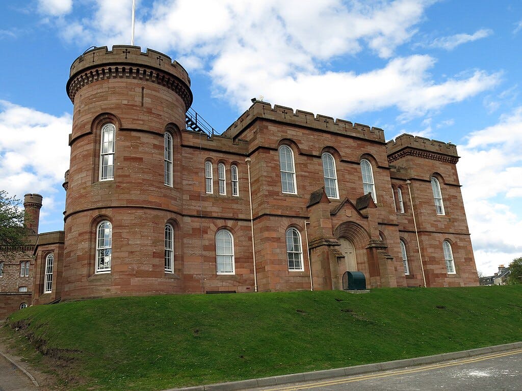 Un edifici de pedra vermella d'estil medieval amb una torre rodona, finestres arquejades i un turó verd de gespa al davant, sota un cel parcialment ennuvolat.