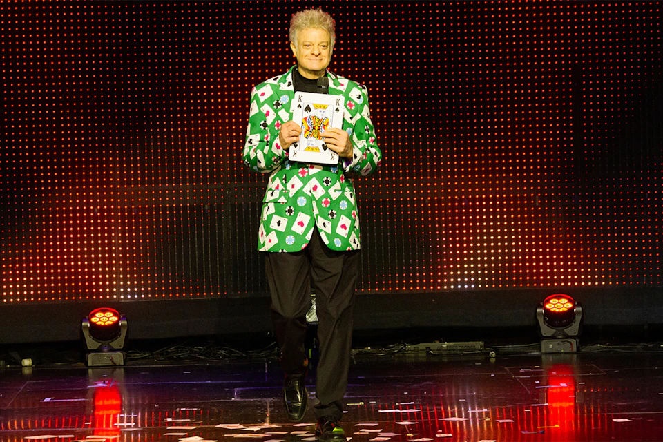 A person wearing a green jacket with card patterns stands on a stage holding a card with a tiger illustration.