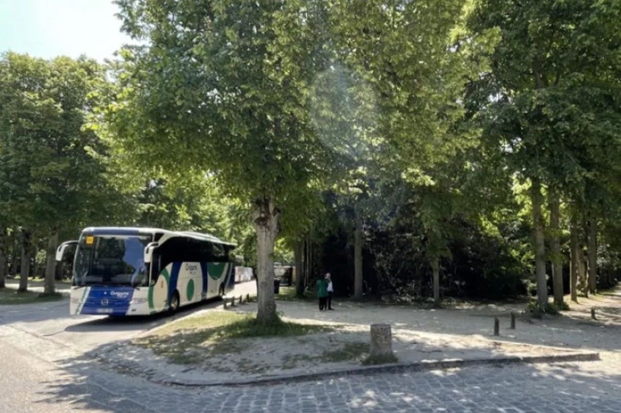 En bus parkeret i nærheden af et træbevokset område med to personer stående i nærheden. Scenen omfatter grønne områder og en asfalteret vej.