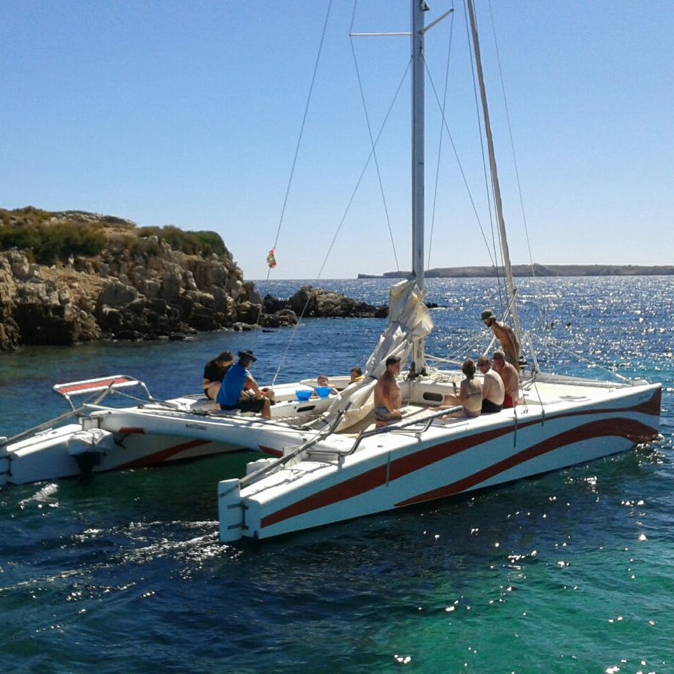 A group of people relax on a white-and-red catamaran in clear blue water near a rocky shoreline under a clear sky.