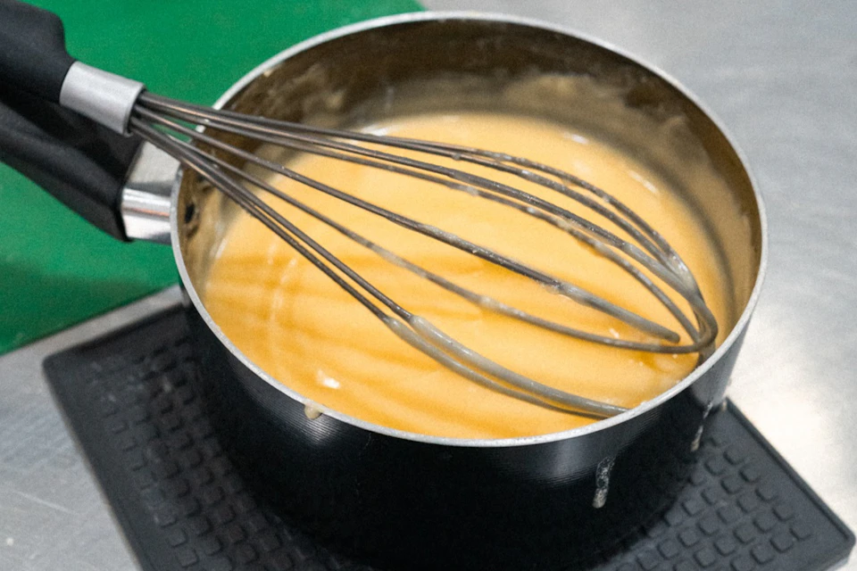 A saucepan filled with a creamy yellow mixture being whisked, placed on a textured surface in a kitchen setting.