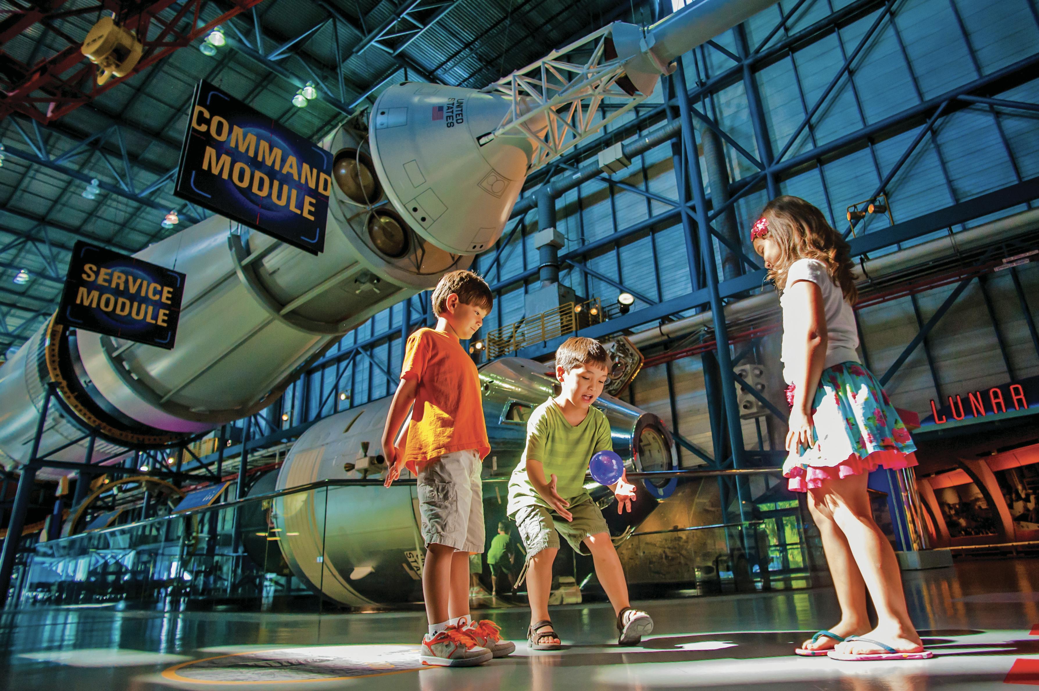 Three children play near a rocket exhibit inside a museum, with a command module displayed above them.