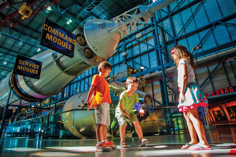 Three children play near a rocket exhibit inside a museum, with a command module displayed above them.