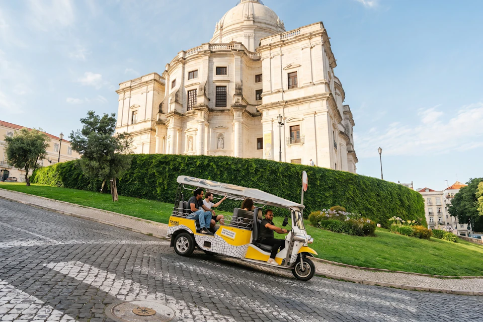 A tuk-tuk with passengers drives on a cobblestone street near a classic, dome-topped building surrounded by greenery.