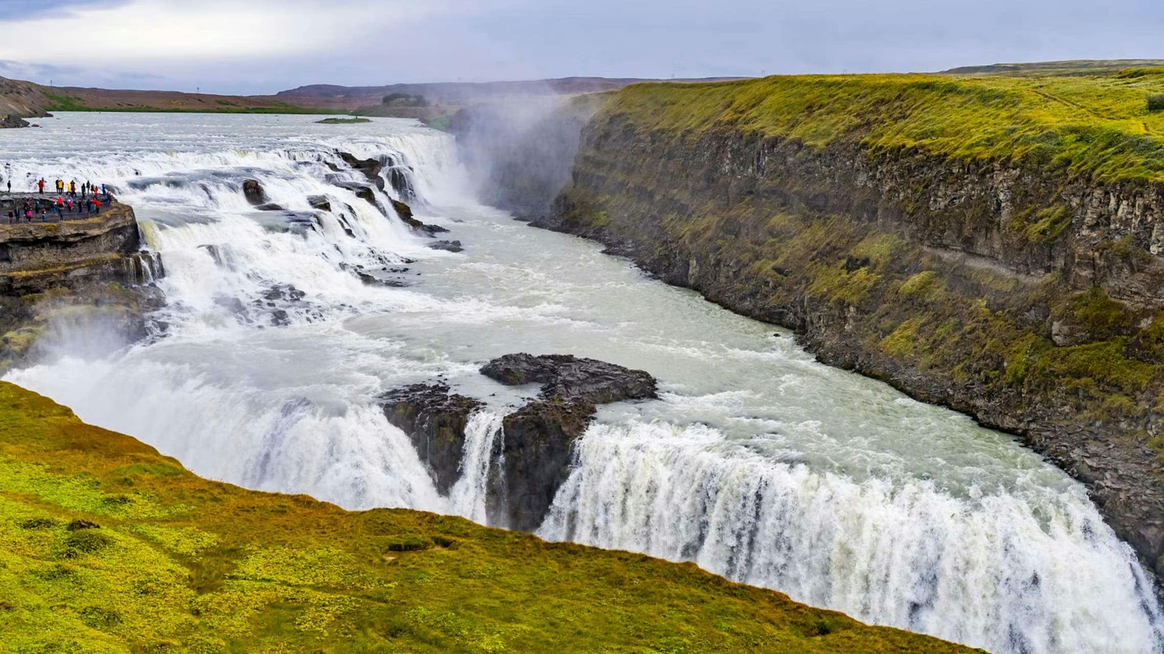 A large waterfall cascades over a cliff into a river below, flanked by grassy, rocky cliffs under a cloudy sky.
