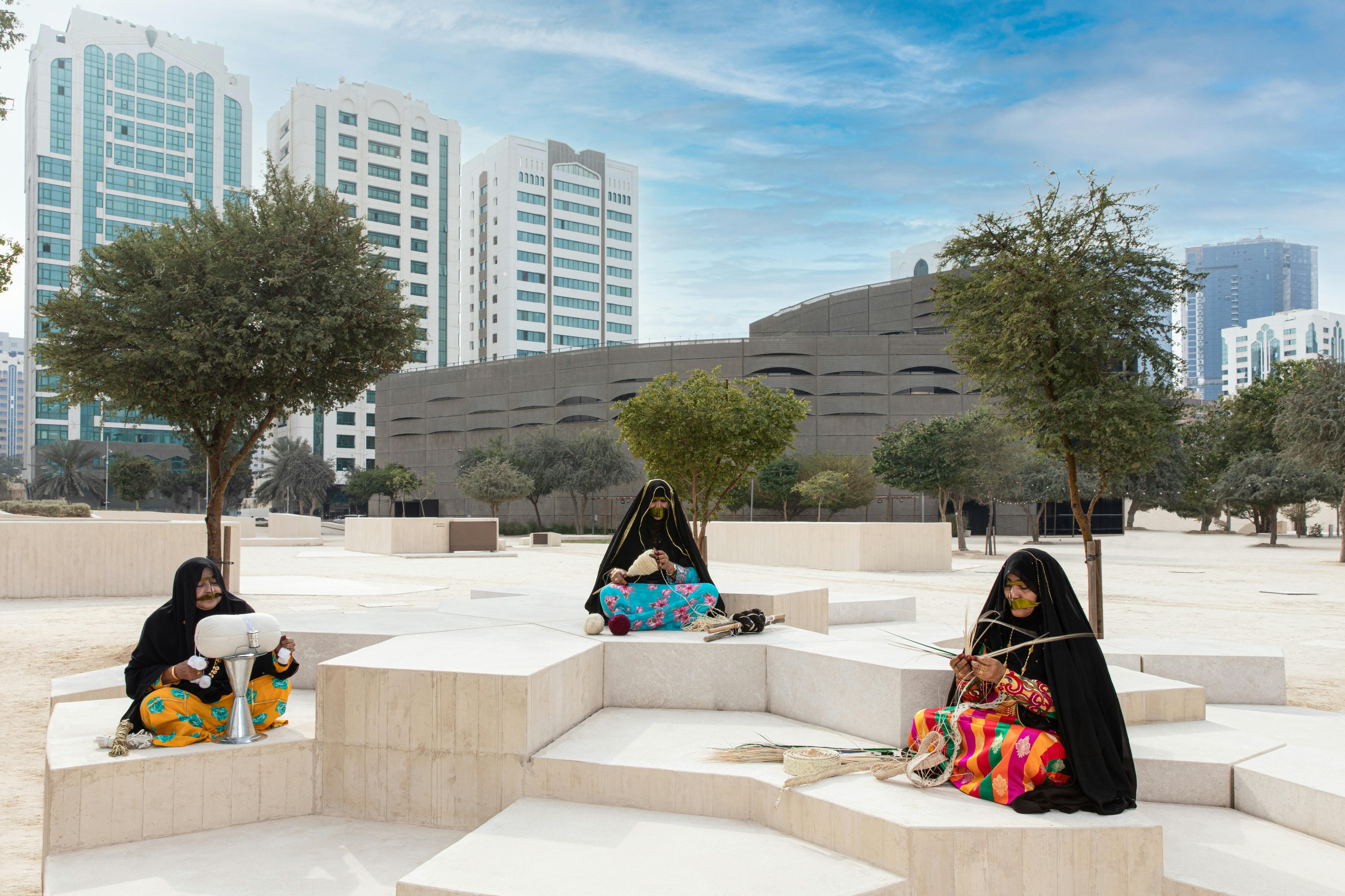 Three women wearing traditional clothing sit on geometric stone structures in a modern urban park with trees and tall buildings in the background.