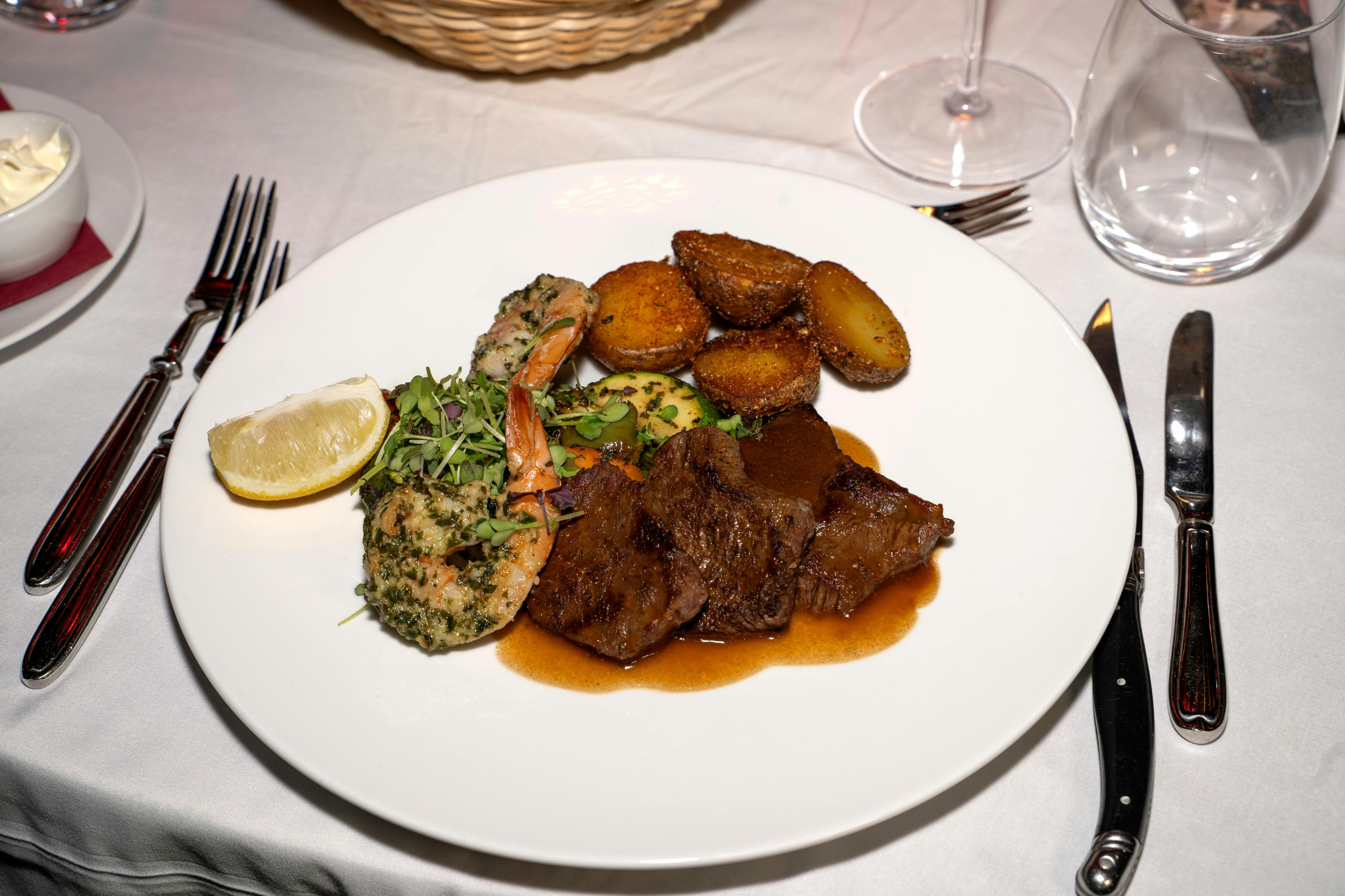 Ένα πιάτο με καρυκευμένες γαρίδες, μπριζόλα, ψητές πατάτες, χόρτα και φέτα λεμονιού, σε λευκό τραπεζομάντιλο με μαχαιροπίρουνα.