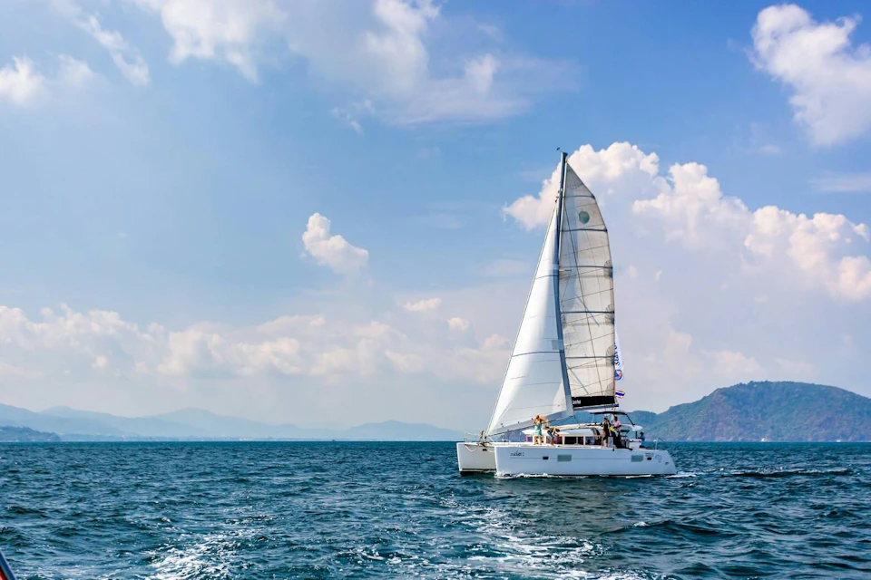 A white sailboat with raised sails on a calm sea under a partly cloudy sky, with distant mountains in the background.