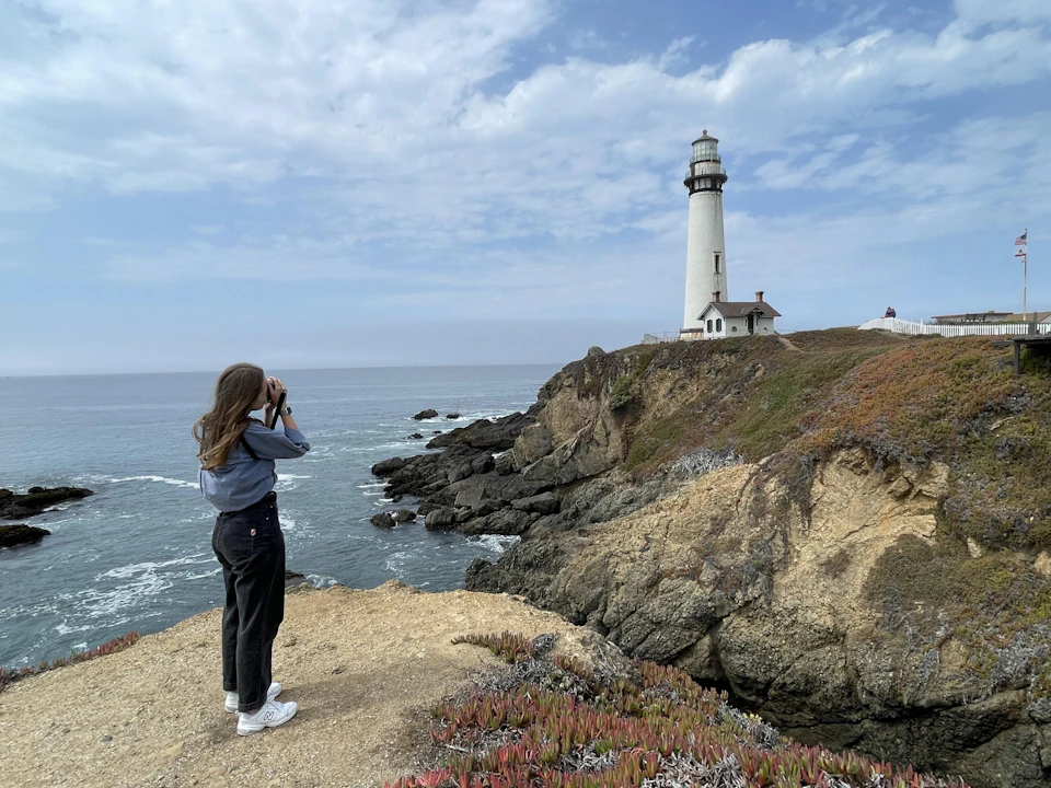 A person with long hair and a camera looks at a lighthouse on a rocky coastal cliff under a partially cloudy sky.