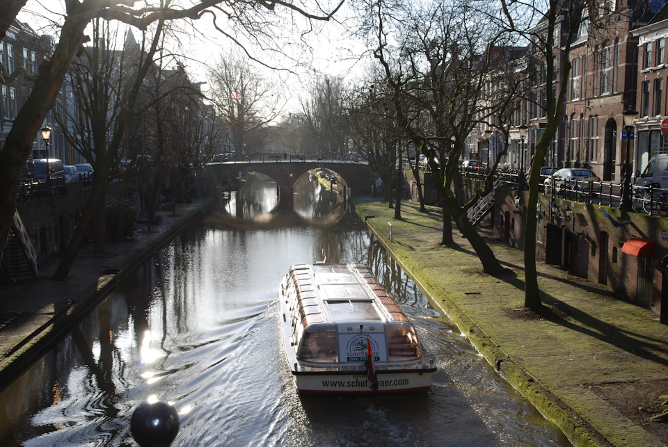A canal boat cruises through a tree-lined waterway toward a stone bridge on a sunny day.
