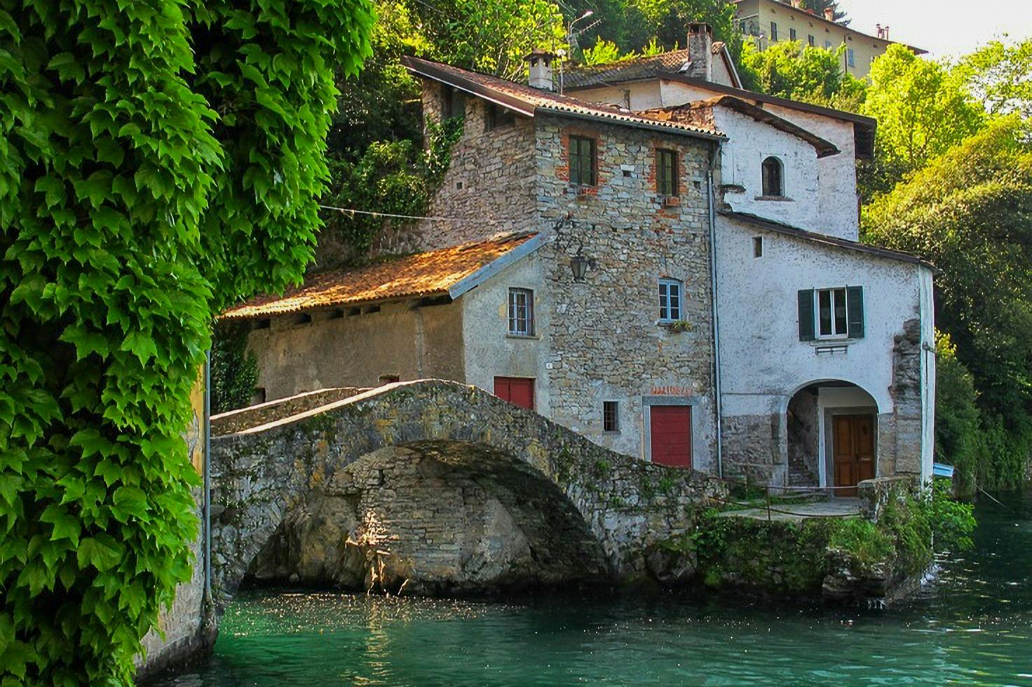 A stone bridge crossing over a calm canal, with rustic stone houses and greenery in the background.