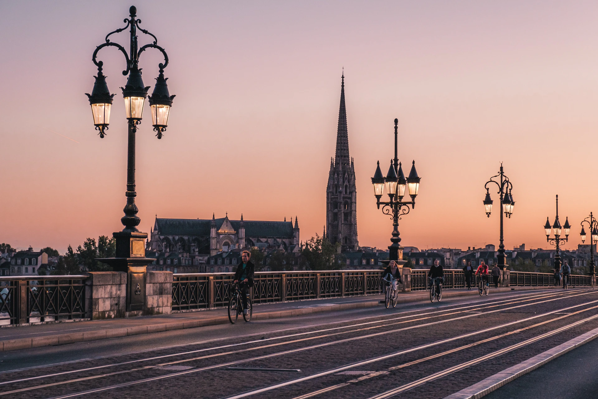 Cyclists ride on a bridge with ornate street lamps at sunset, with a tall historic spire and a church in the background.