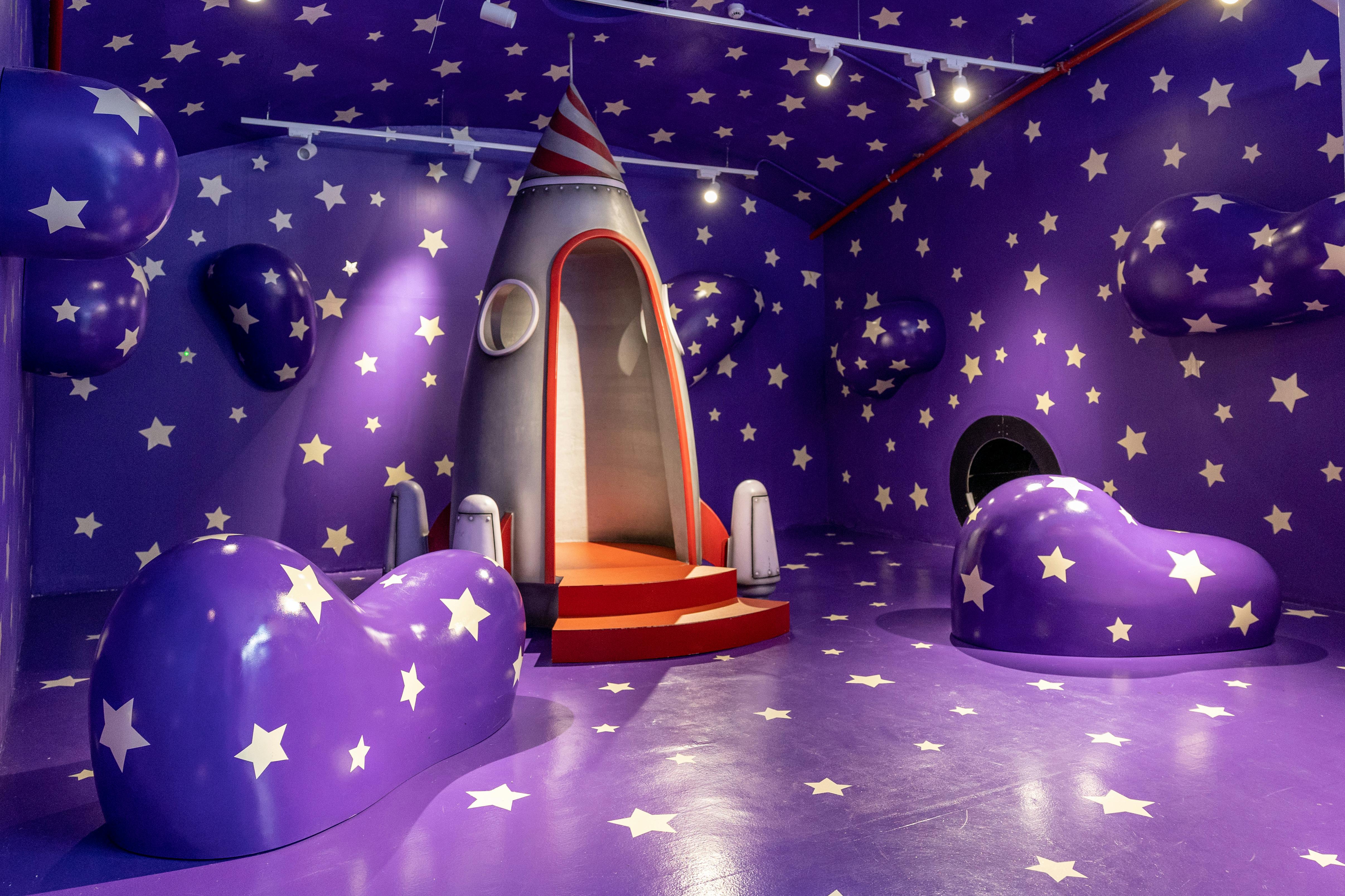 Room with purple walls and floor covered in white stars, featuring a central rocket play structure and curved seating.