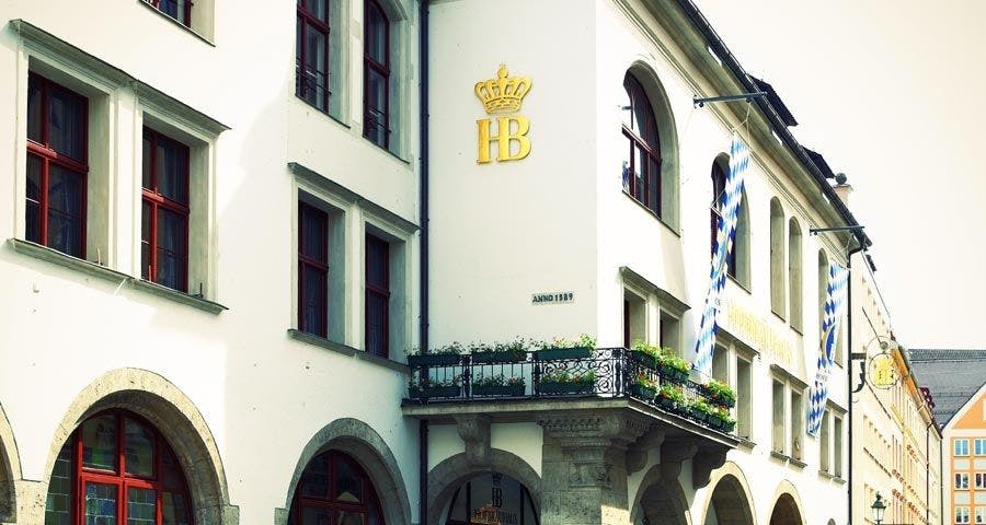 Historická budova s klenutými okny, dekorativním balkonem a logem zlaté koruny s nápisem „HB“ na fasádě. Banner a rostliny přítomny.