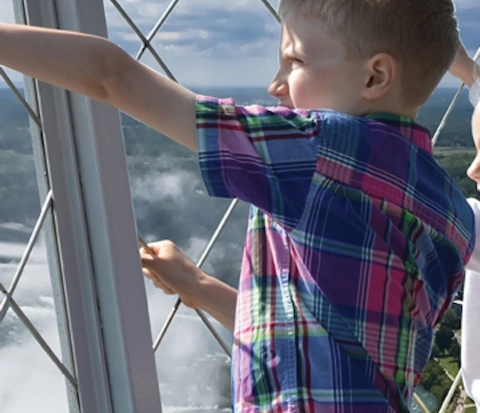 Two children excitedly point out the window at a large waterfall below, framed by a metal lattice structure.