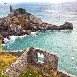 En kyrka och gamla stenmurar på en stenig kustlinje med utsikt över blågrönt vatten med en avlägsen båt vid horisonten.