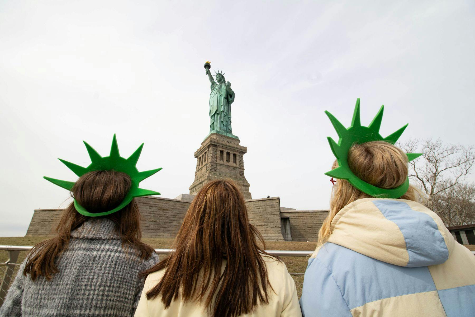 Drie mensen met groene Statue of Liberty kroonhoeden op kijken uit op het Vrijheidsbeeld op een bewolkte dag.