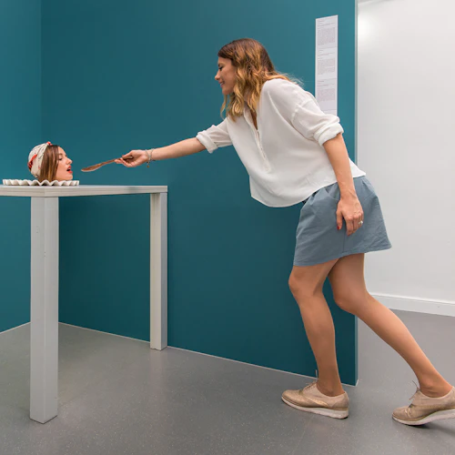 En kvinna matar ett disembodied huvud på ett bord med en träsked; en blomstervas är bredvid huvudet.