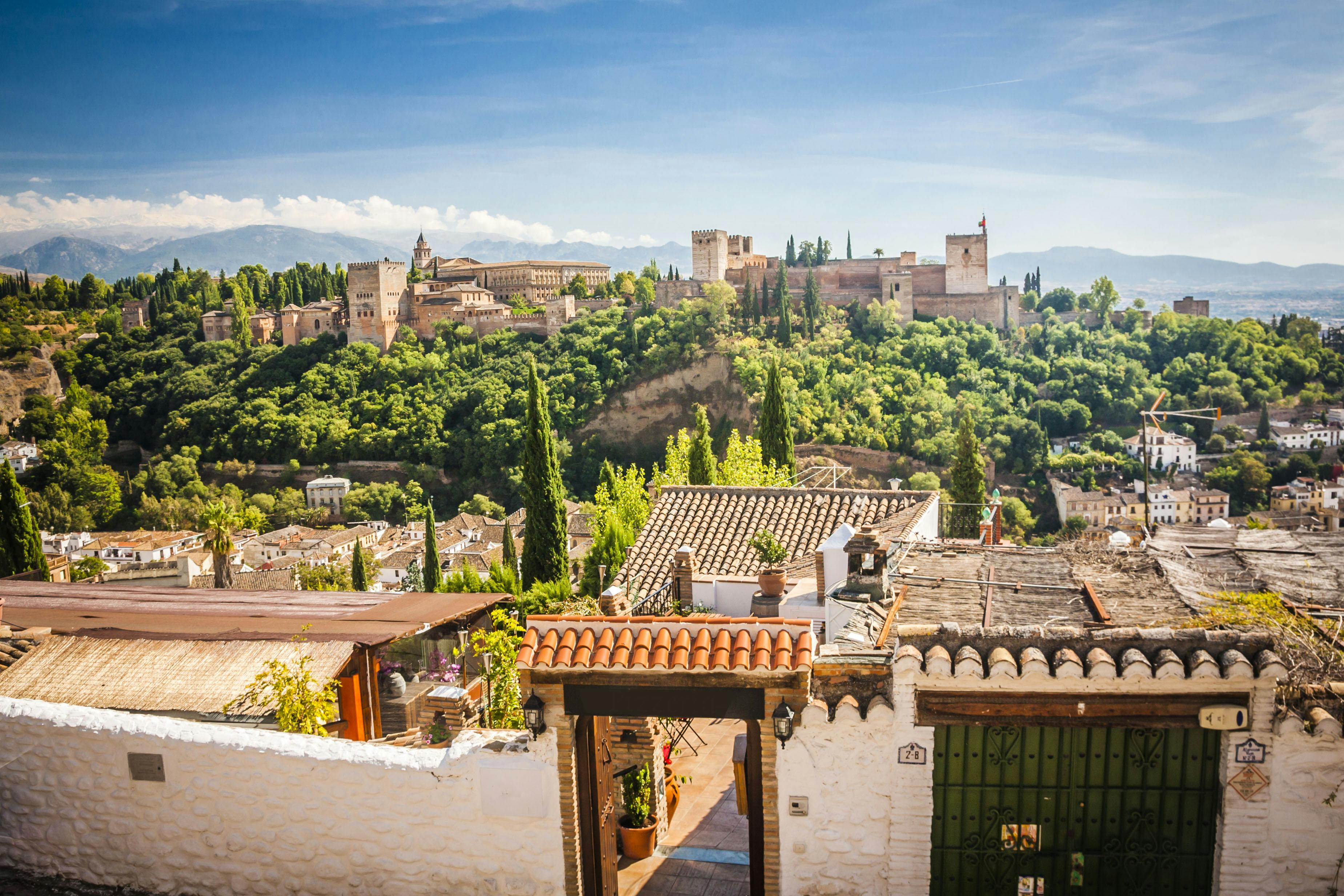 A hilltop fortress surrounded by lush greenery, with a foreground of tiled rooftops and white buildings under a clear sky.