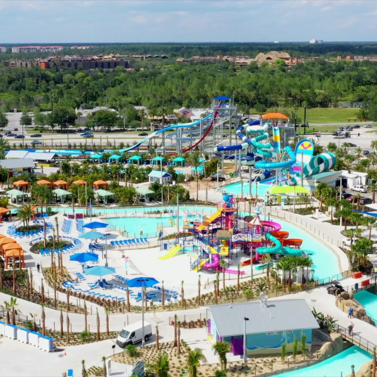 Aerial view of a waterpark with various colorful slides, pools, and lounging areas surrounded by trees and parking.