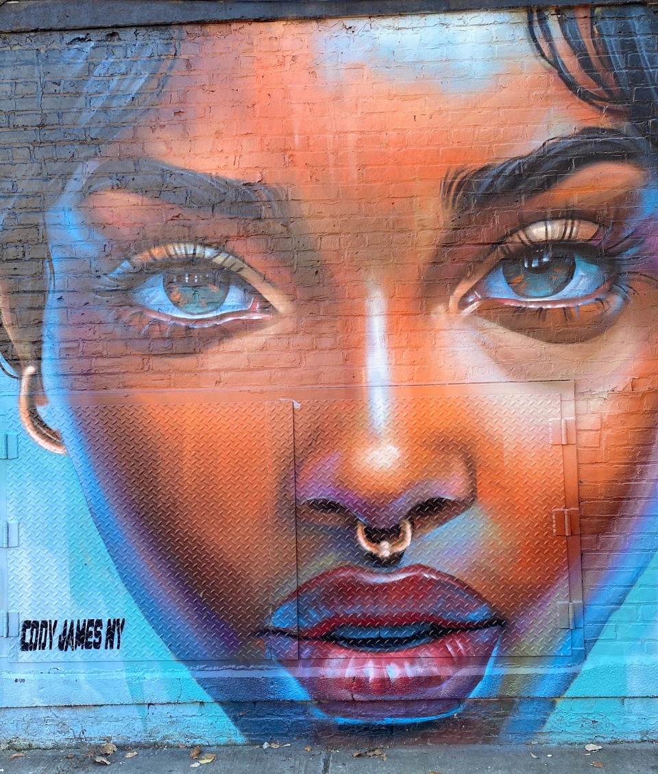 A vibrant mural on a brick wall depicting a close-up of a woman's face with expressive eyes, a nose ring, and colorful tones.