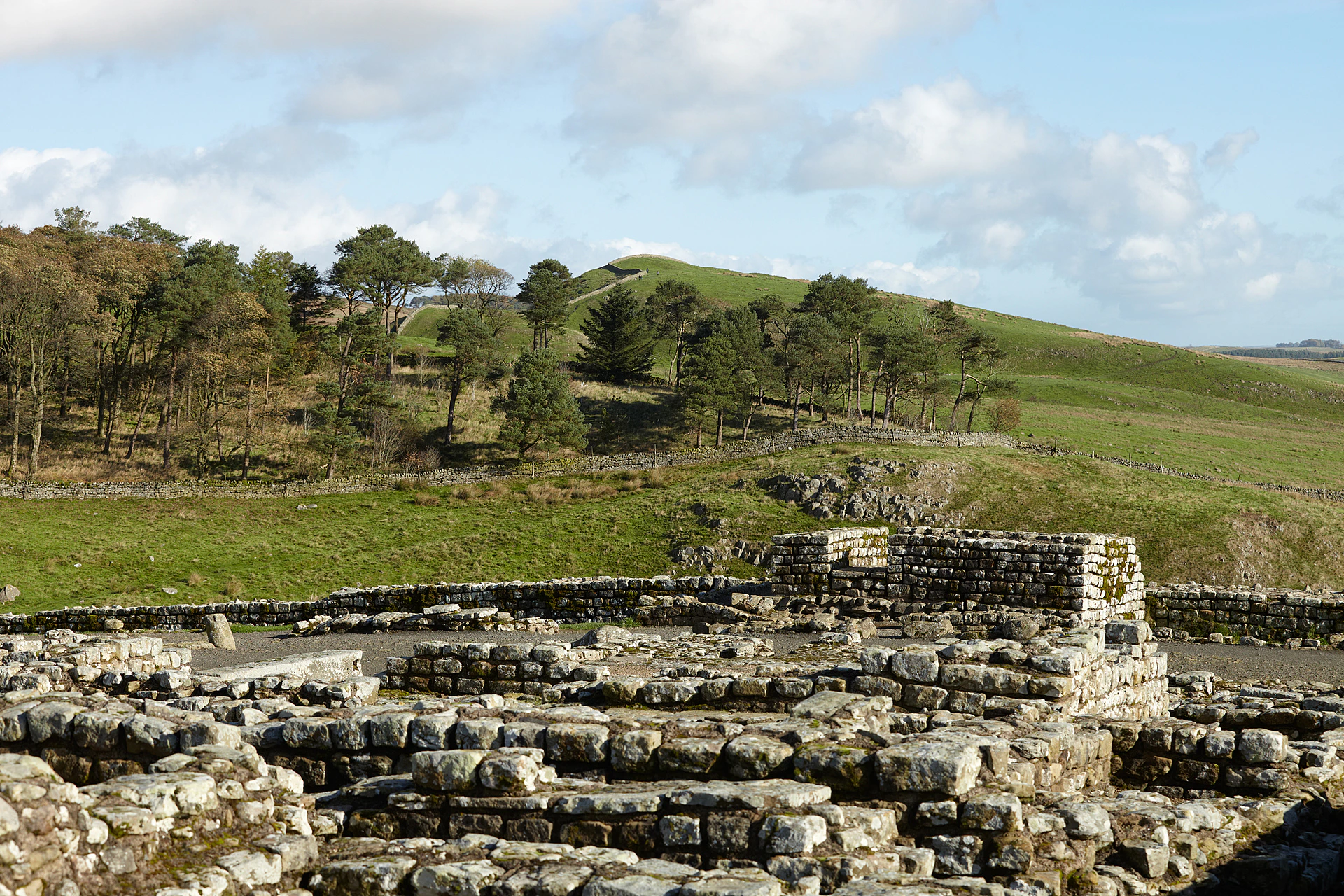 Ancient stone ruins in the foreground with green rolling hills and trees under a partly cloudy sky in the background.