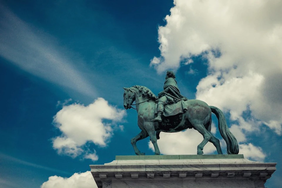 Bronze equestrian statue of a robed figure on a pedestal against a blue sky with scattered clouds.
