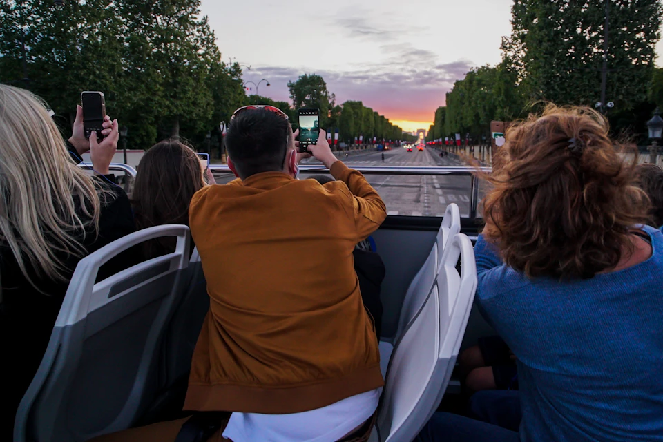 A group of people sitting on an open-top bus, taking photos of a sunset over a tree-lined street.
