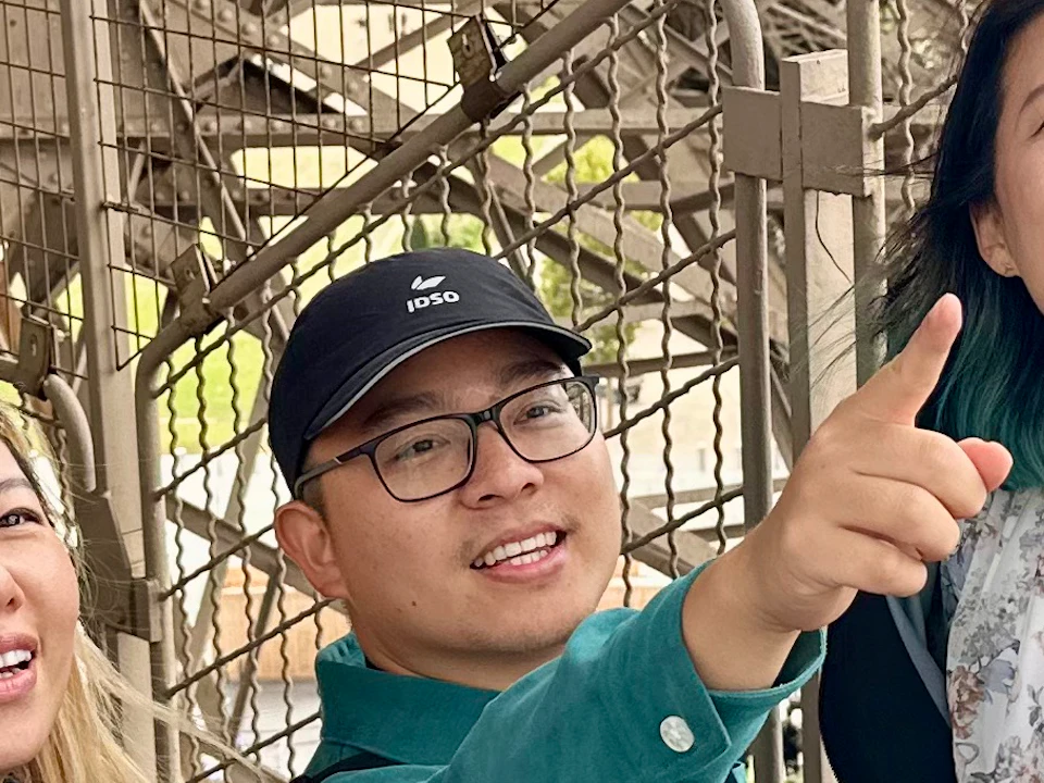 Three people smile and point while standing inside the Eiffel Tower's structure, with metal beams and mesh fencing around them.