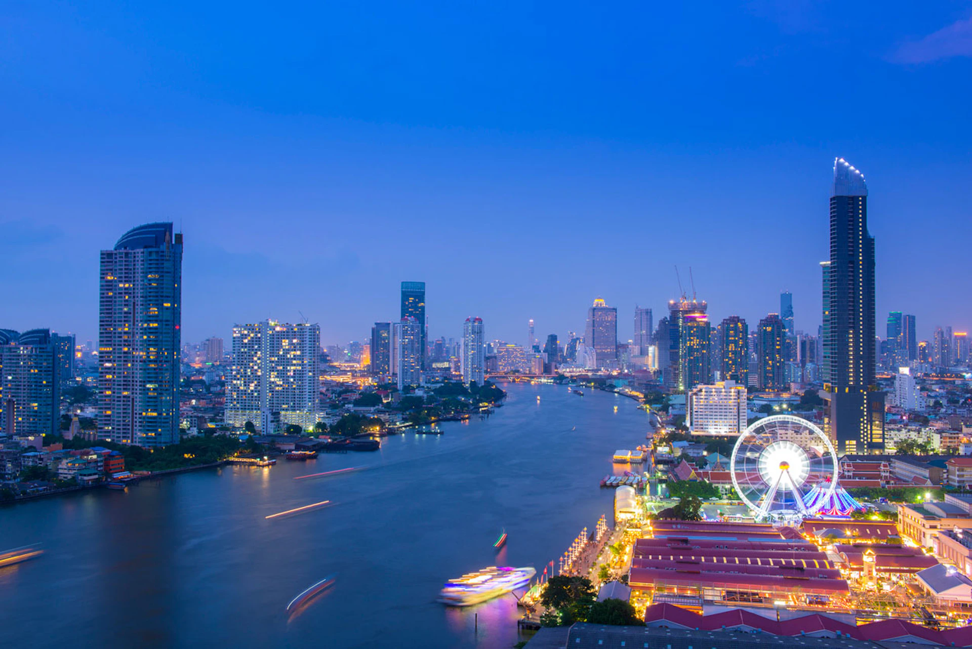 Cityscape at dusk featuring a river, illuminated buildings, a colorful riverfront, and a large Ferris wheel.