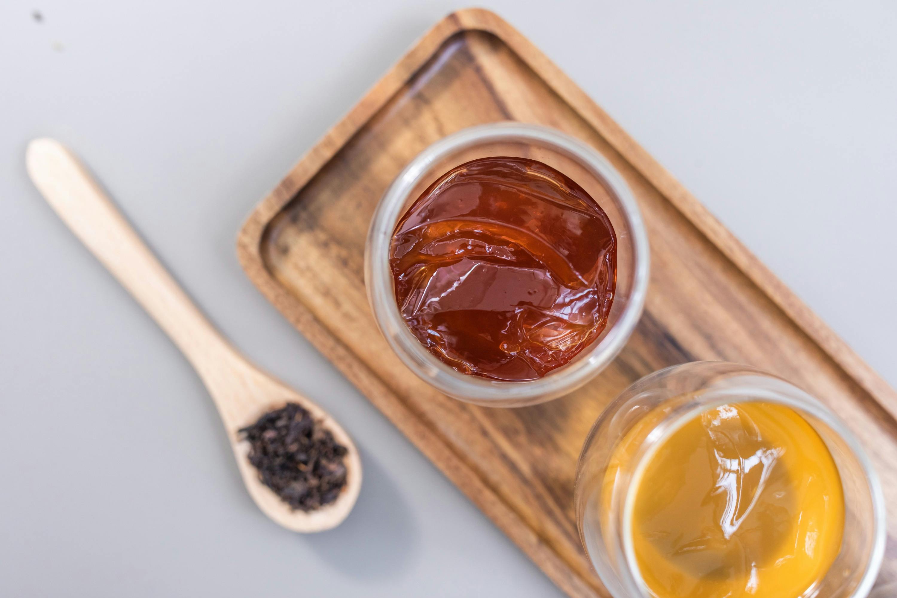 Two glass cups of tea, one dark and one light, on a wooden tray, next to a wooden spoon with loose tea leaves.