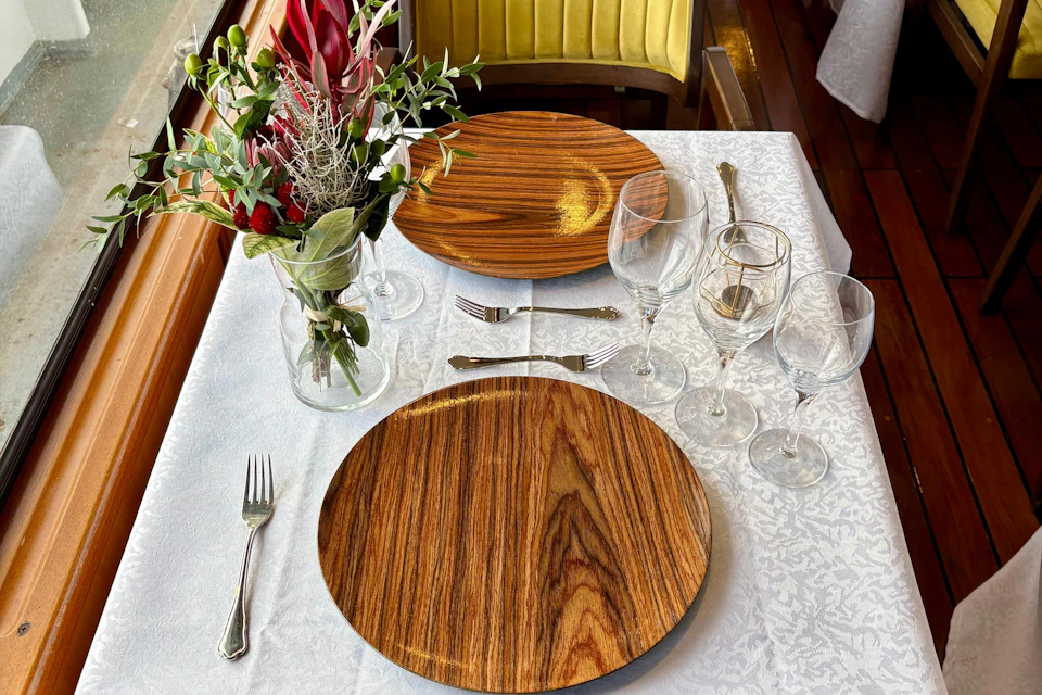 A table set for two with wooden plates, wine glasses, and cutlery on a white tablecloth, featuring a floral arrangement centerpiece.
