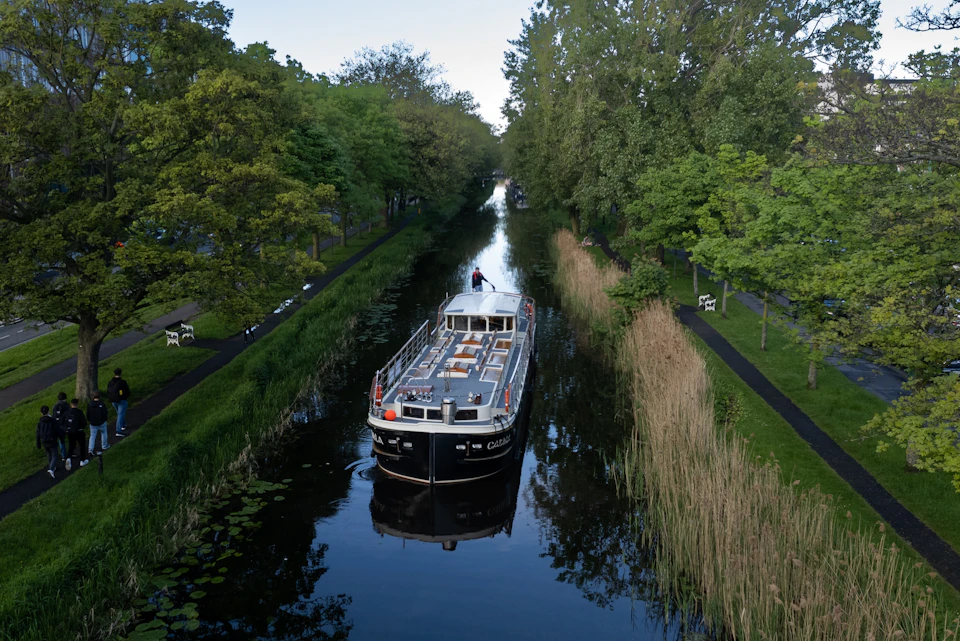 A boat travels through a narrow canal surrounded by lush green trees and reeds under a clear sky.