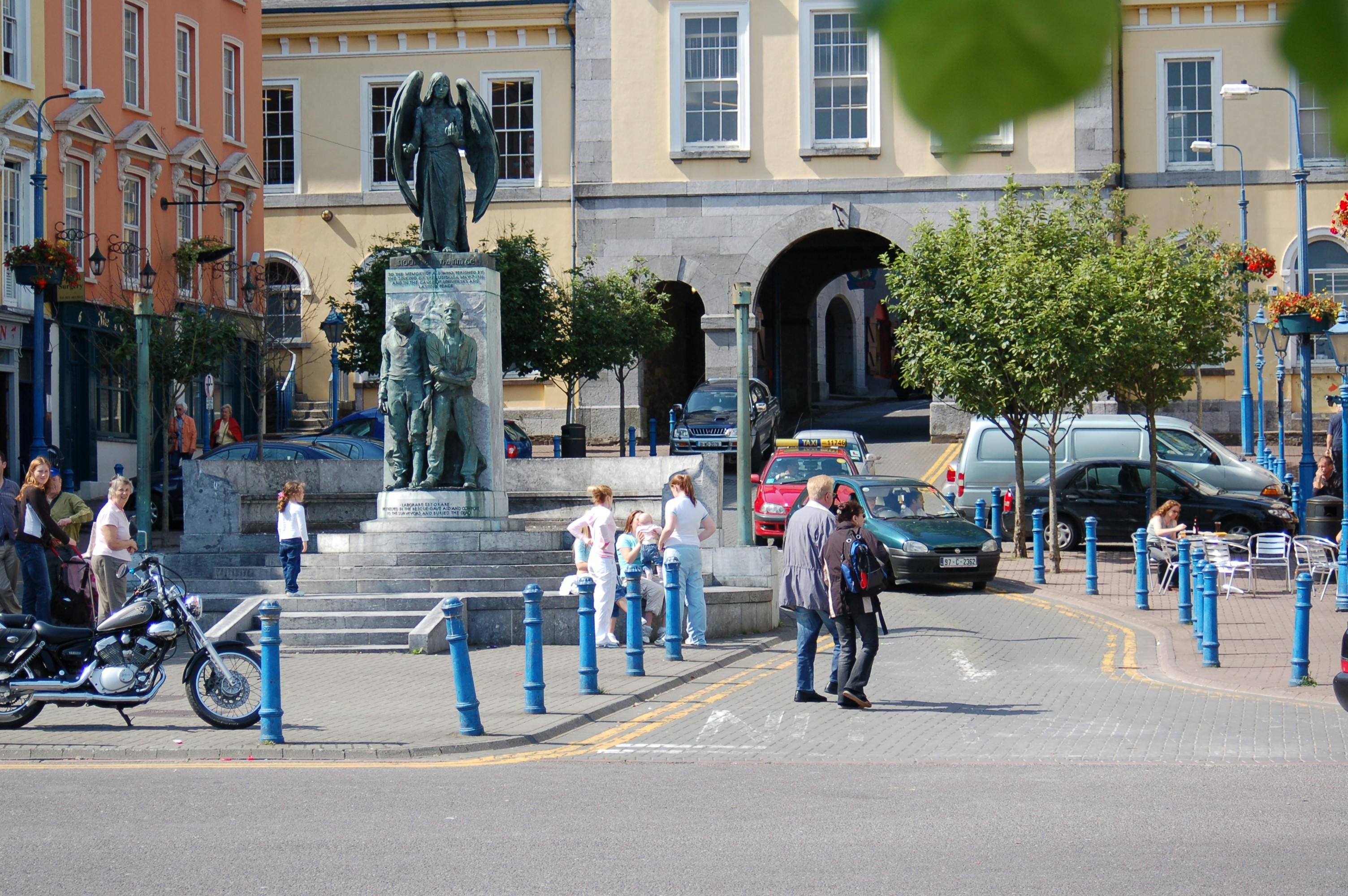 A town square features a bronze war memorial statue with people walking, a motorcycle, and colorful buildings in the background.