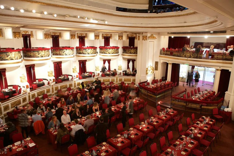 A grand dining hall with red tablecloths, chandeliers, and two levels of seating. People are dining and conversing.
