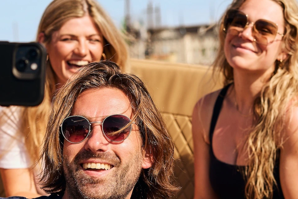 A group of people smiling and taking a selfie while sitting outdoors on a sunny day, holding glasses and enjoying themselves.