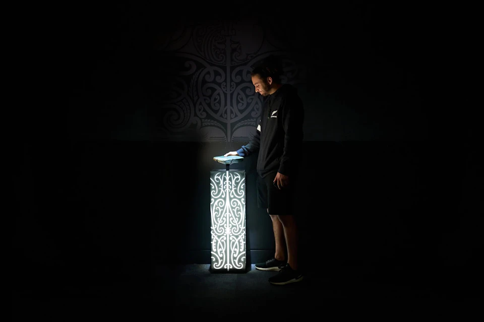 A person interacts with a glowing pedestal featuring intricate patterns, set against a dark background with faint designs.