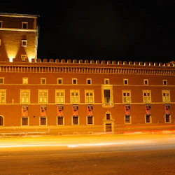 En upplyst historisk byggnad med banderoller på fasaden på natten, med ljusspår från passerande fordon i förgrunden.