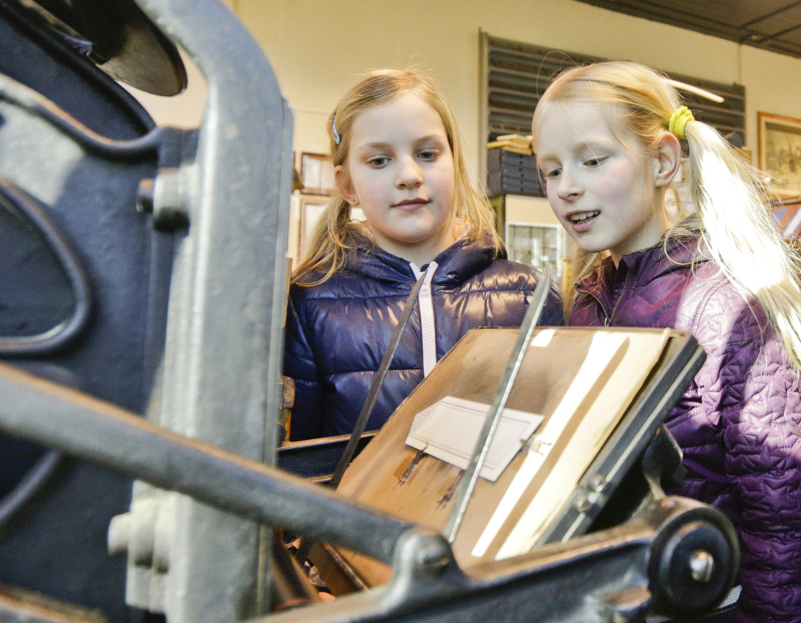 Две молодые девушки, обе в фиолетовых куртках, наблюдают за винтажным печатным станком в хорошо освещенном помещении.