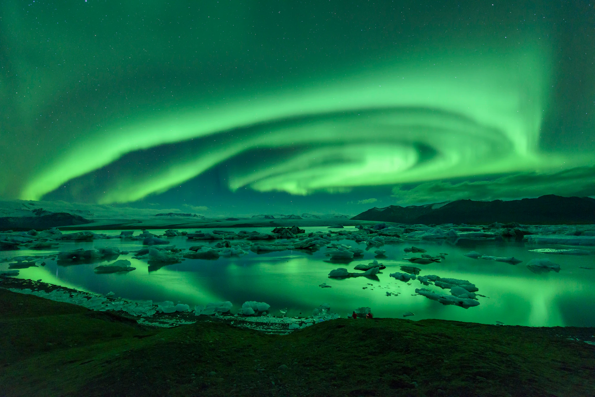 The Northern Lights illuminate a night sky over an icy landscape with a reflective lake and scattered icebergs.