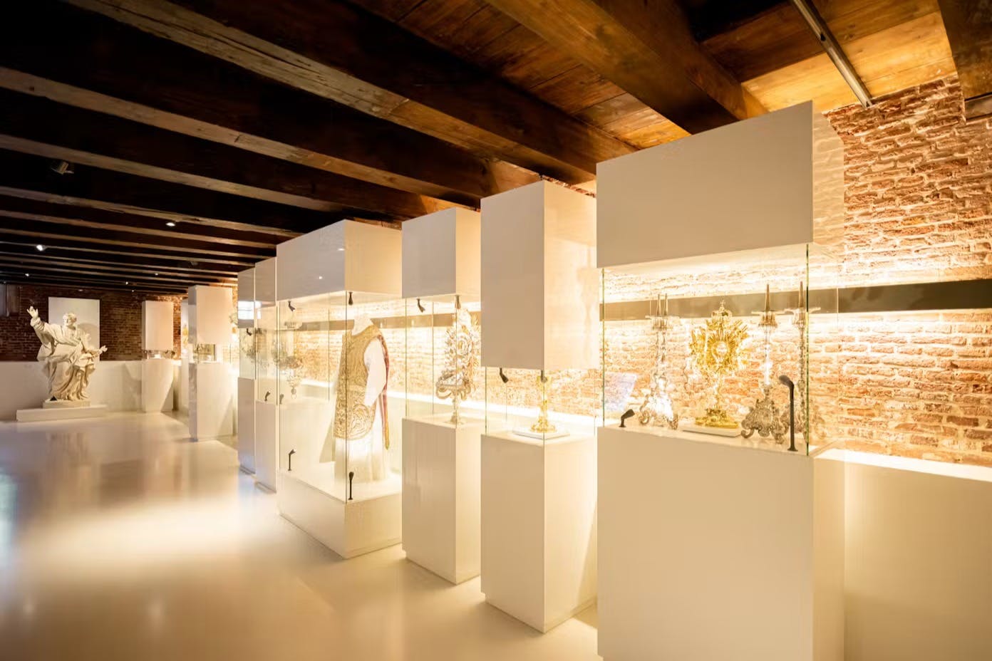 Exposição num museu moderno, com vitrinas de vidro que contêm artefactos e peças de vestuário ornamentados, sob um teto com vigas de madeira.