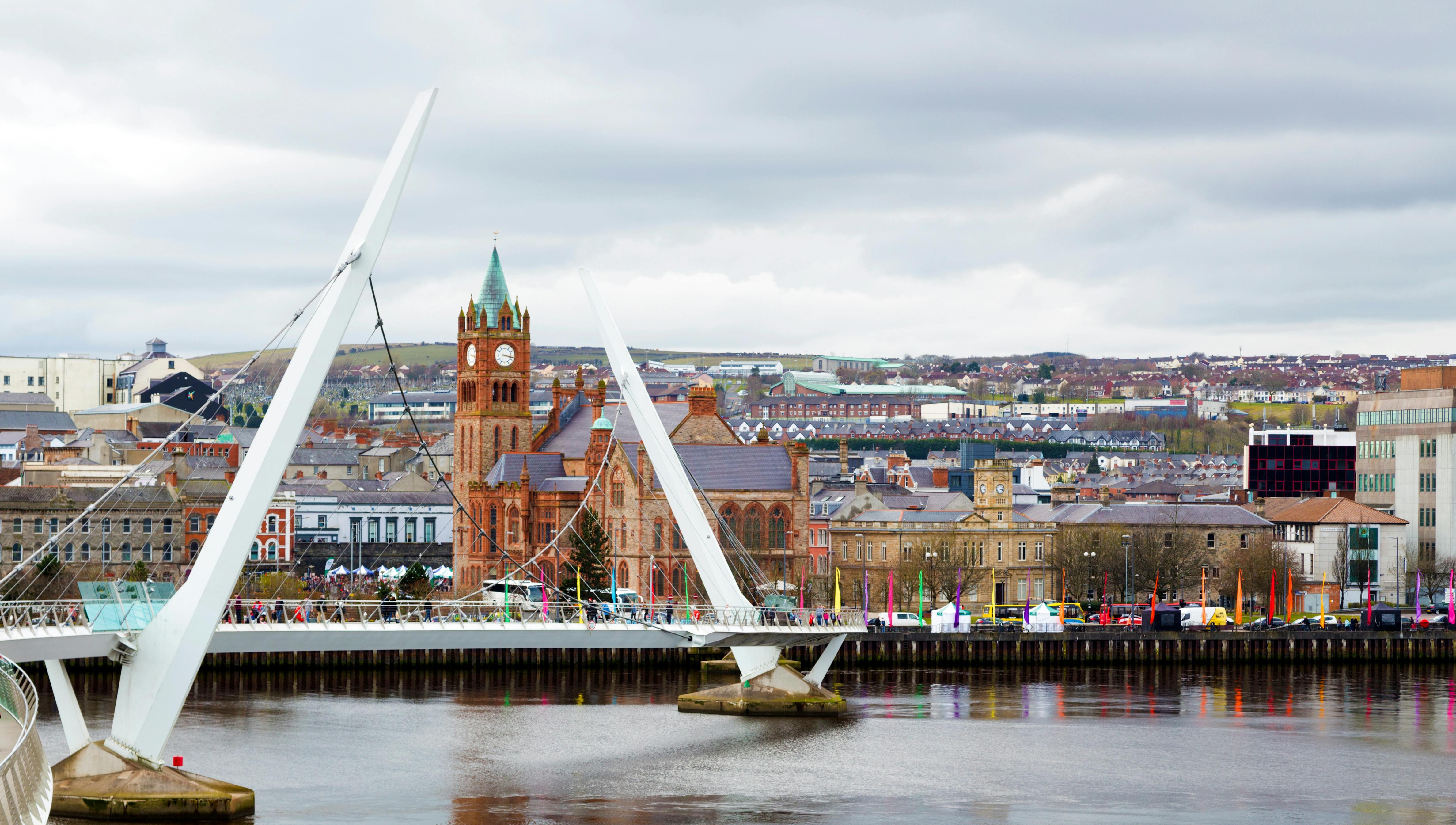 Modern bridge over a river with people walking, a historic clock tower, and buildings in the background under a cloudy sky.