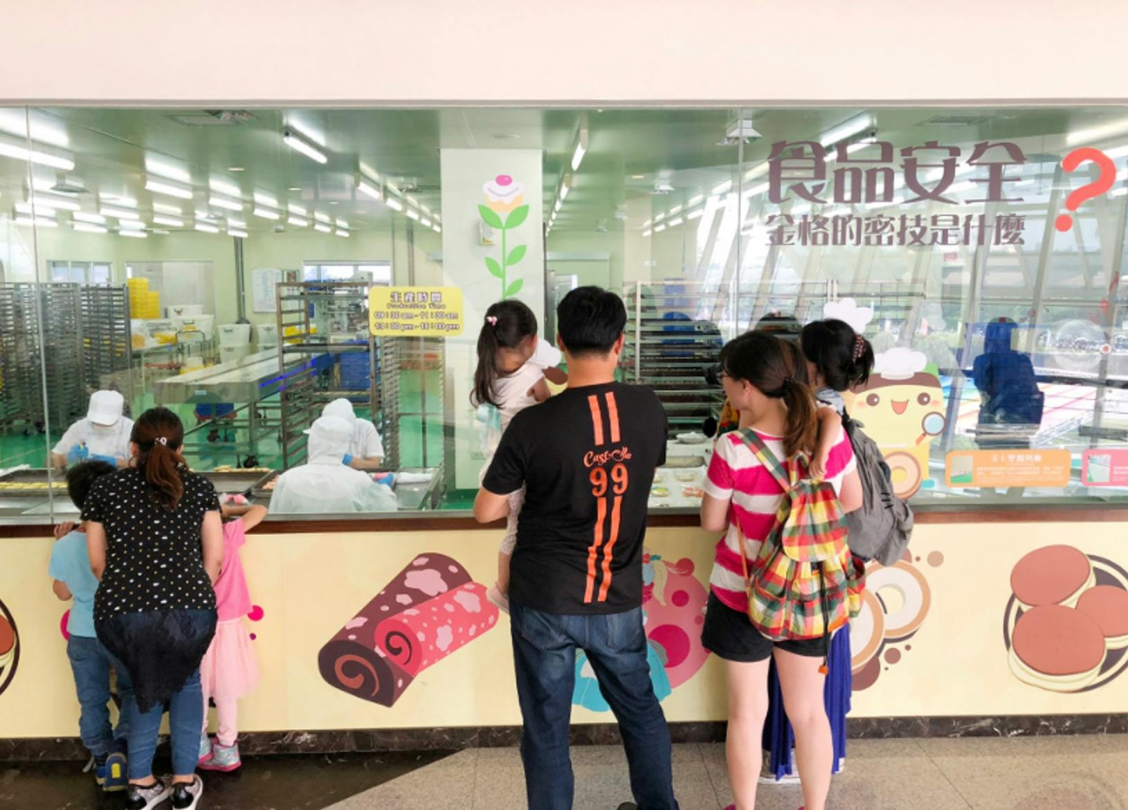A group of people, including a child, looking into a bakery production area through a glass window with colorful decorations.