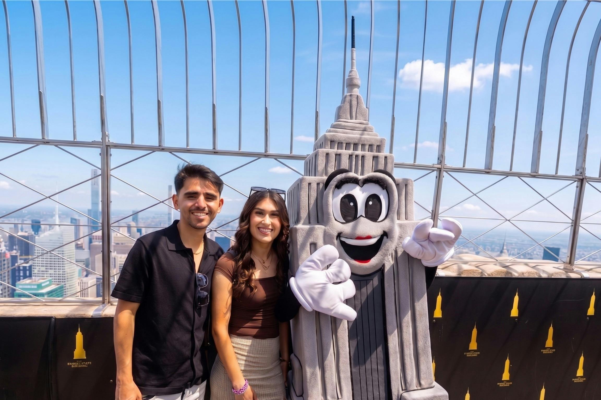 Twee mensen die naast iemand in een Empire State Building-kostuum staan op een uitkijkplatform, met op de achtergrond het uitzicht over de stad.