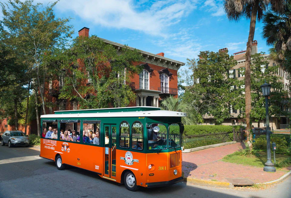 An orange-and-green tour trolley moves past a historic red-brick building adorned with greenery. Passengers are visible inside.